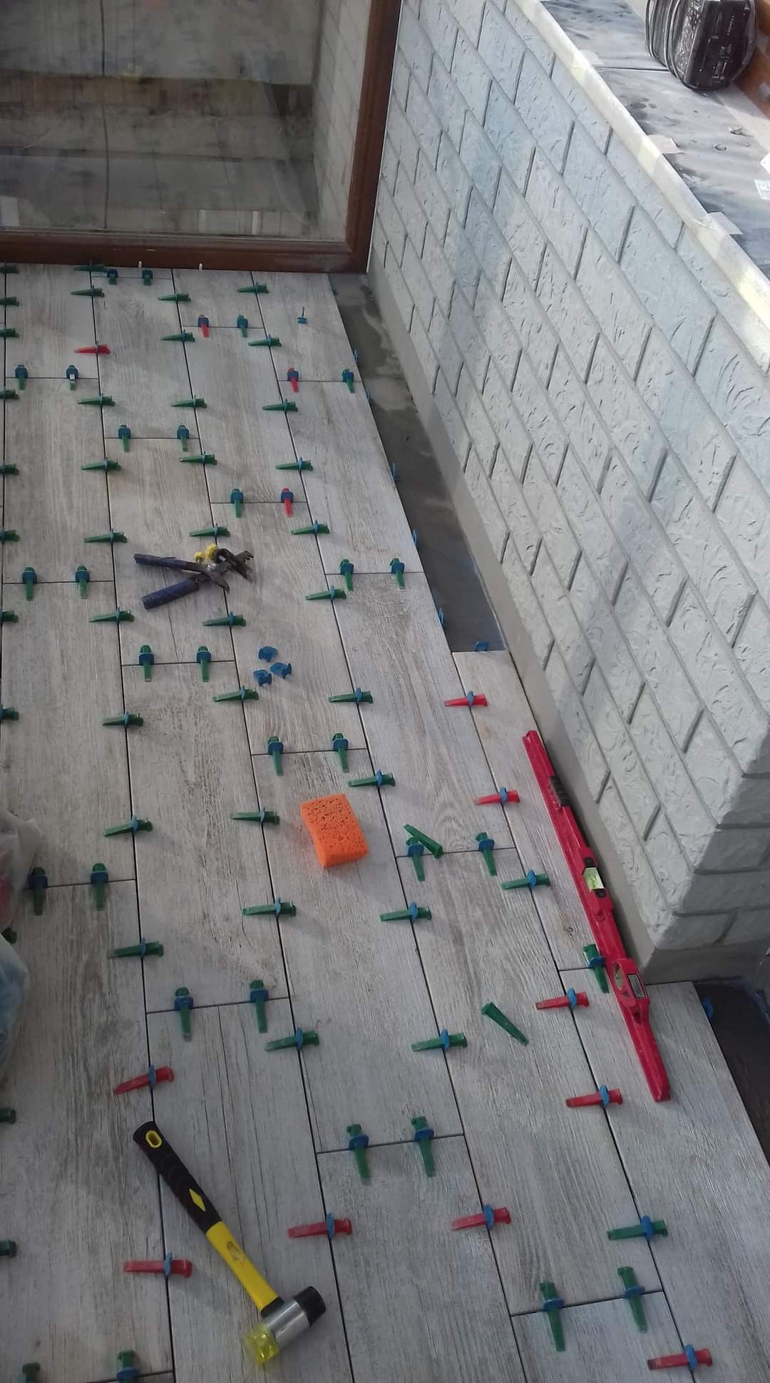 Układanie jasnych płytek podłogowych z użyciem systemu poziomowania, widoczny poziomica, młotek gumowy i szczypce do płytek.