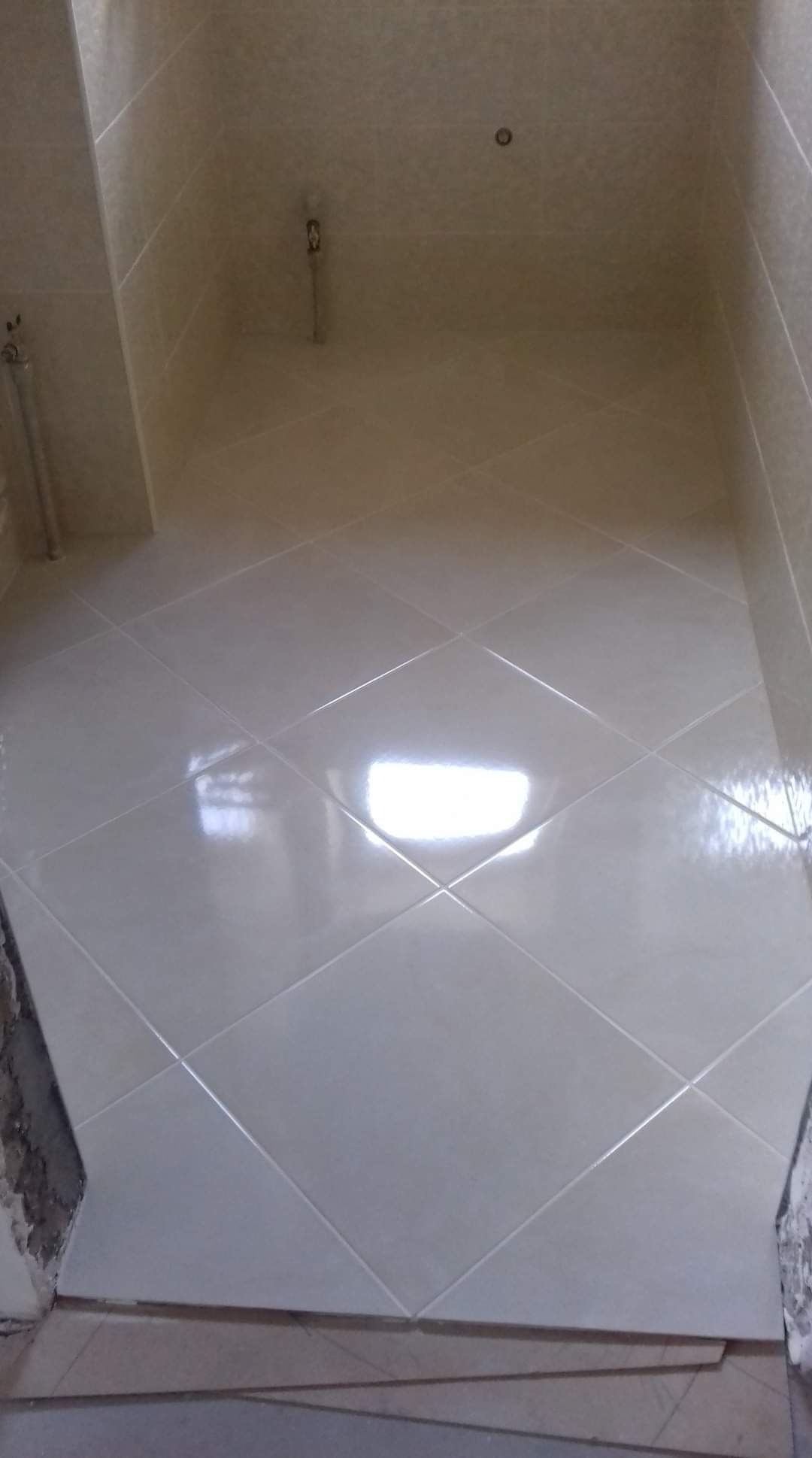 Wykończona łazienka z jasnymi, błyszczącymi płytkami ułożonymi pod kątem, widoczne pozostałości po pracach wykończeniowych przy podłodze.