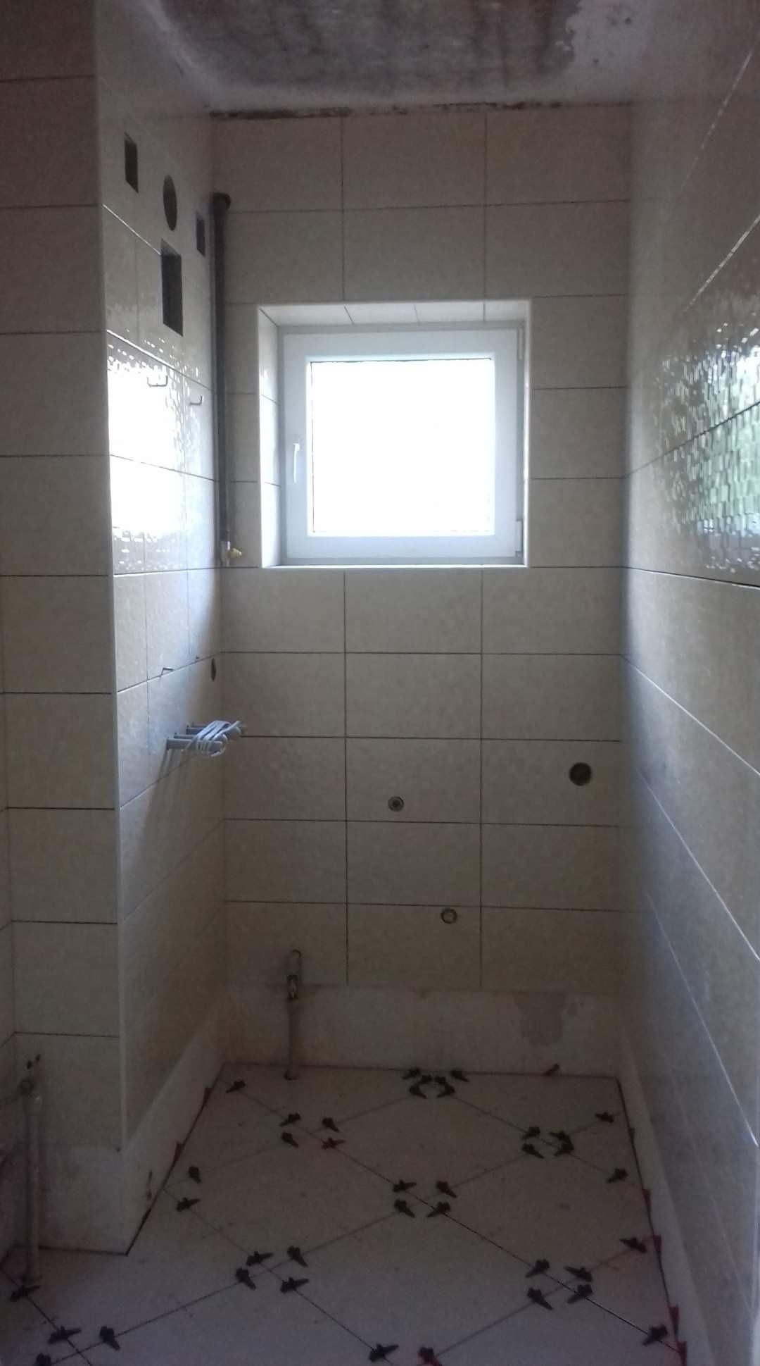 Łazienka w trakcie remontu: ułożone płytki podłogowe z krzyżykami dystansowymi, częściowo wykończone ściany z jasnymi płytkami, widoczne rury i otwory instalacyjne.