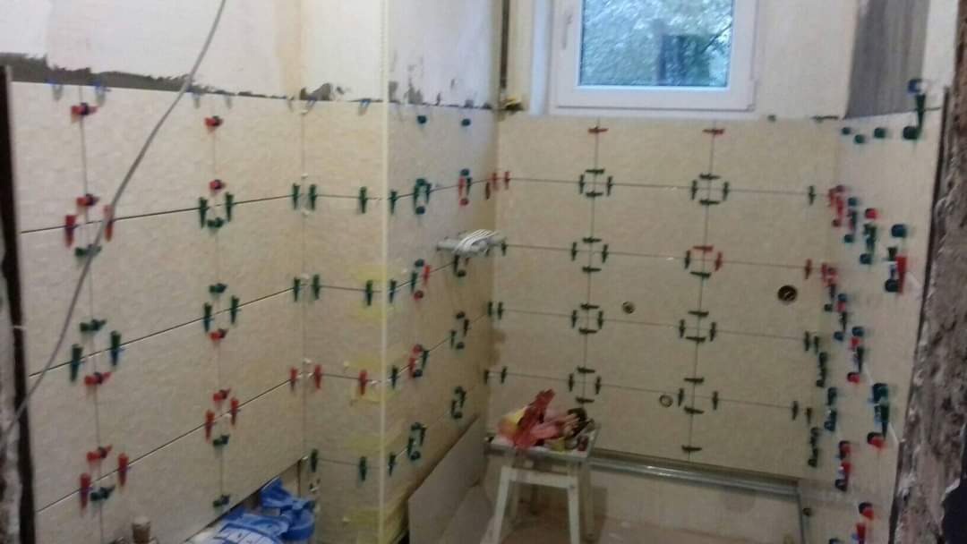 Układanie jasnych płytek na ścianie w łazience, widoczne kolorowe krzyżyki dystansowe i poziomica laserowa.