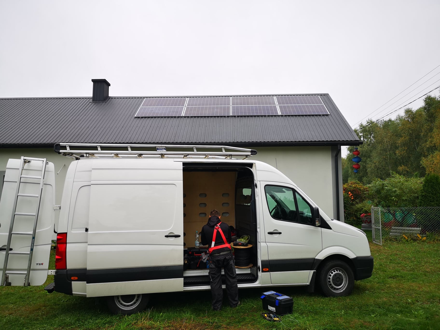 Pracownik w szelkach bezpieczeństwa przy otwartym vanie serwisowym na tle domu z panelami słonecznymi na dachu.