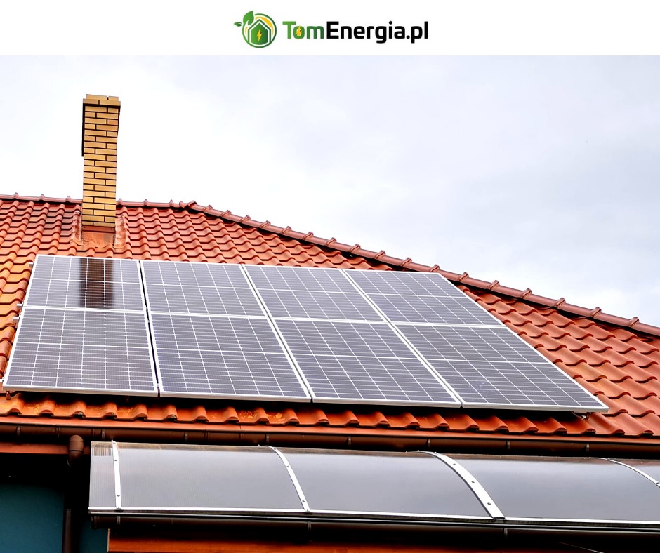 Panele słoneczne zamontowane na dachu z czerwonej dachówki, widoczny komin z cegły i logo TomEnergia.pl w górnym rogu.