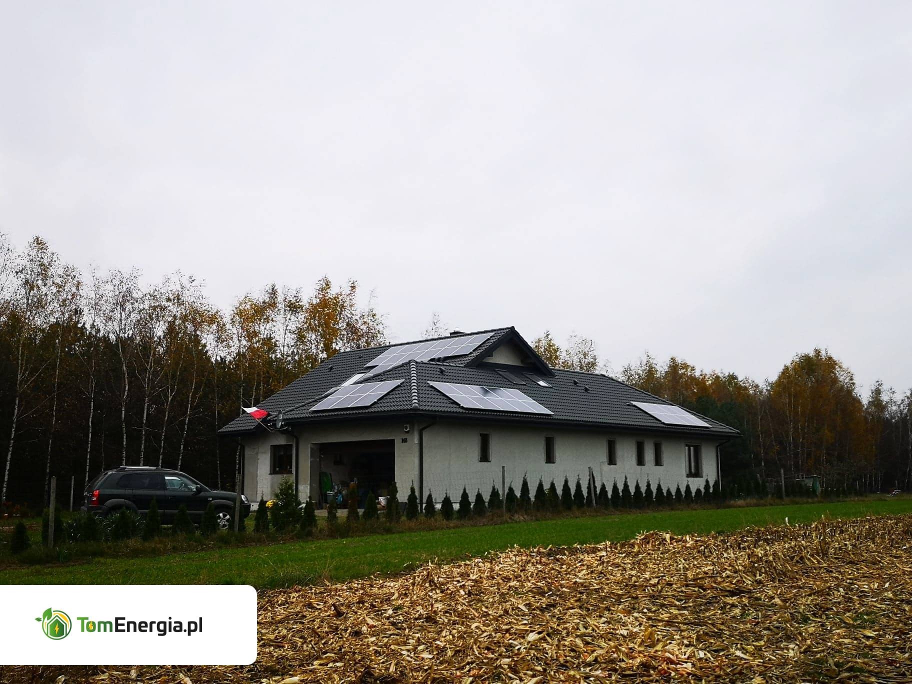 Dom jednorodzinny z zamontowanymi panelami słonecznymi na dachu, widok z zewnątrz, z samochodem zaparkowanym obok i polem kukurydzy na pierwszym planie.