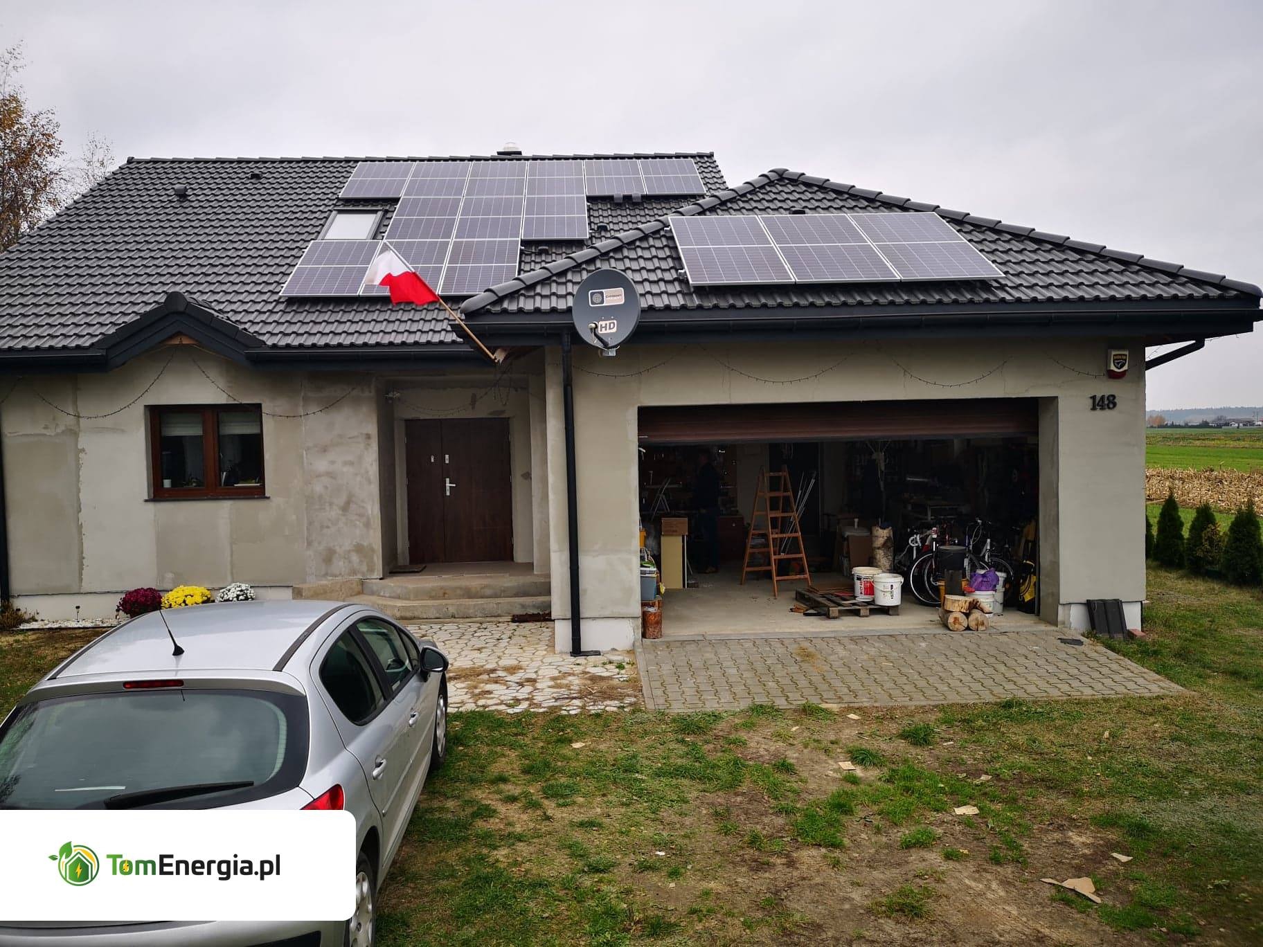 Dom jednorodzinny z panelami fotowoltaicznymi na dachu, udekorowany polską flagą, garaż z otwartą bramą, na podjeździe srebrny samochód osobowy.