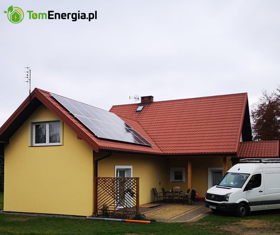 Żółty dom jednorodzinny z panelami słonecznymi na dachu, czerwona dachówka, zaparkowany biały van firmy instalacyjnej, widoczne logo firmy w lewym górnym rogu.