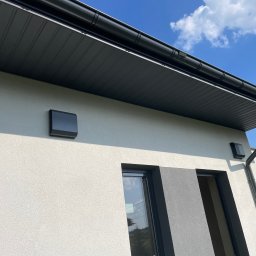 MarKal INSTAL - Elewacja budynku z zamontowanymi czarnymi lampami zewnętrznymi, widoczny fragment okna, drzwi i orynnowanie na tle błękitnego nieba z chmurami.