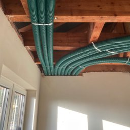 MarKal INSTAL - Zielone, karbowane rury wentylacyjne zamontowane pod drewnianym stropem, przymocowane perforowanymi metalowymi taśmami, widoczne okno i fragment ściany w surowym stanie.