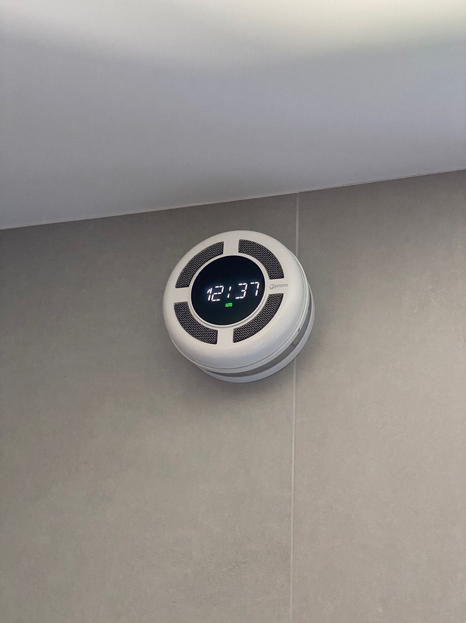 Okrągły, biały rekuperator ścienny z wyświetlaczem cyfrowym wskazującym godzinę 12:37, zamontowany na szarej, gładkiej ścianie.