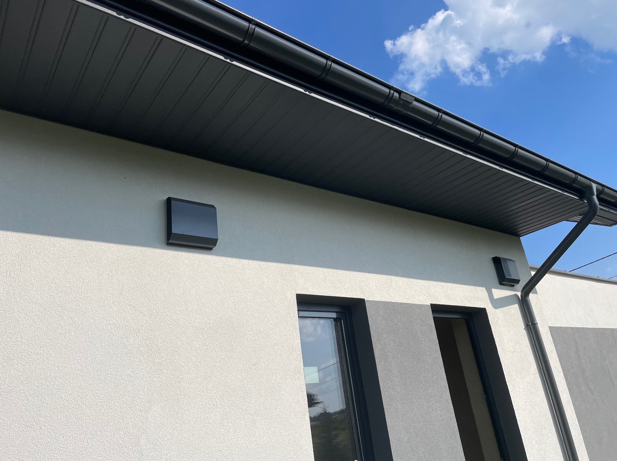 Elewacja budynku z zamontowanymi czarnymi lampami zewnętrznymi, widoczny fragment okna, drzwi i orynnowanie na tle błękitnego nieba z chmurami.