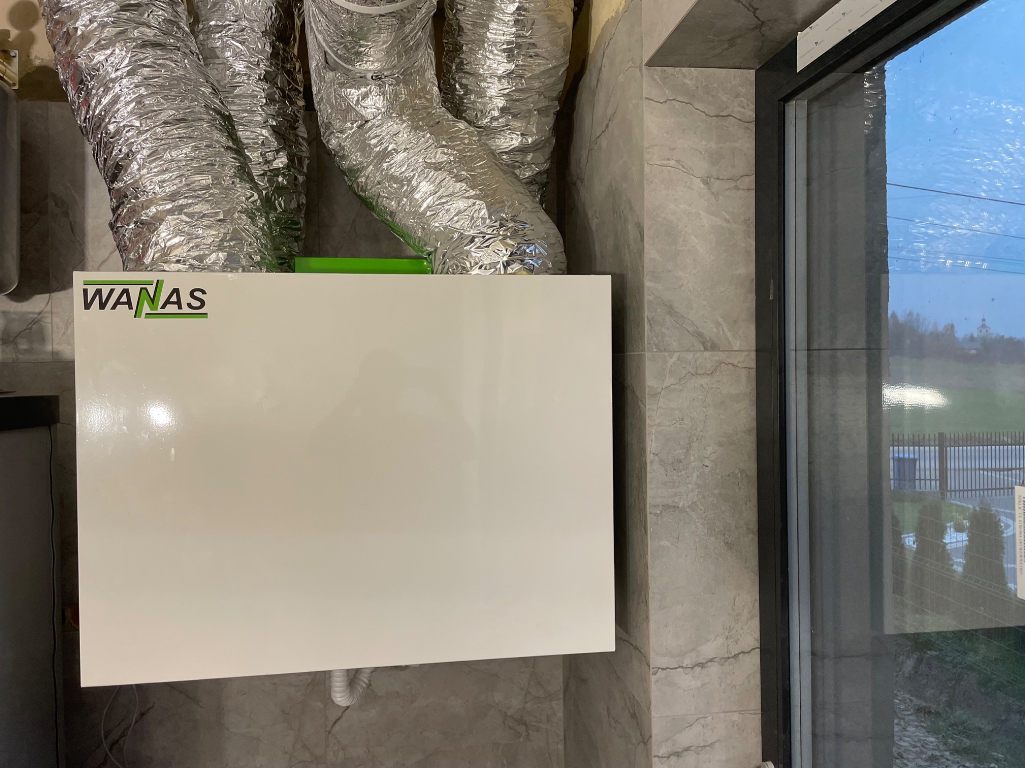 Biała jednostka rekuperacji marki WANAS z podłączonymi srebrnymi przewodami wentylacyjnymi, zamontowana na ścianie z szarych płytek imitujących marmur, widok przez okno na zewnątrz.