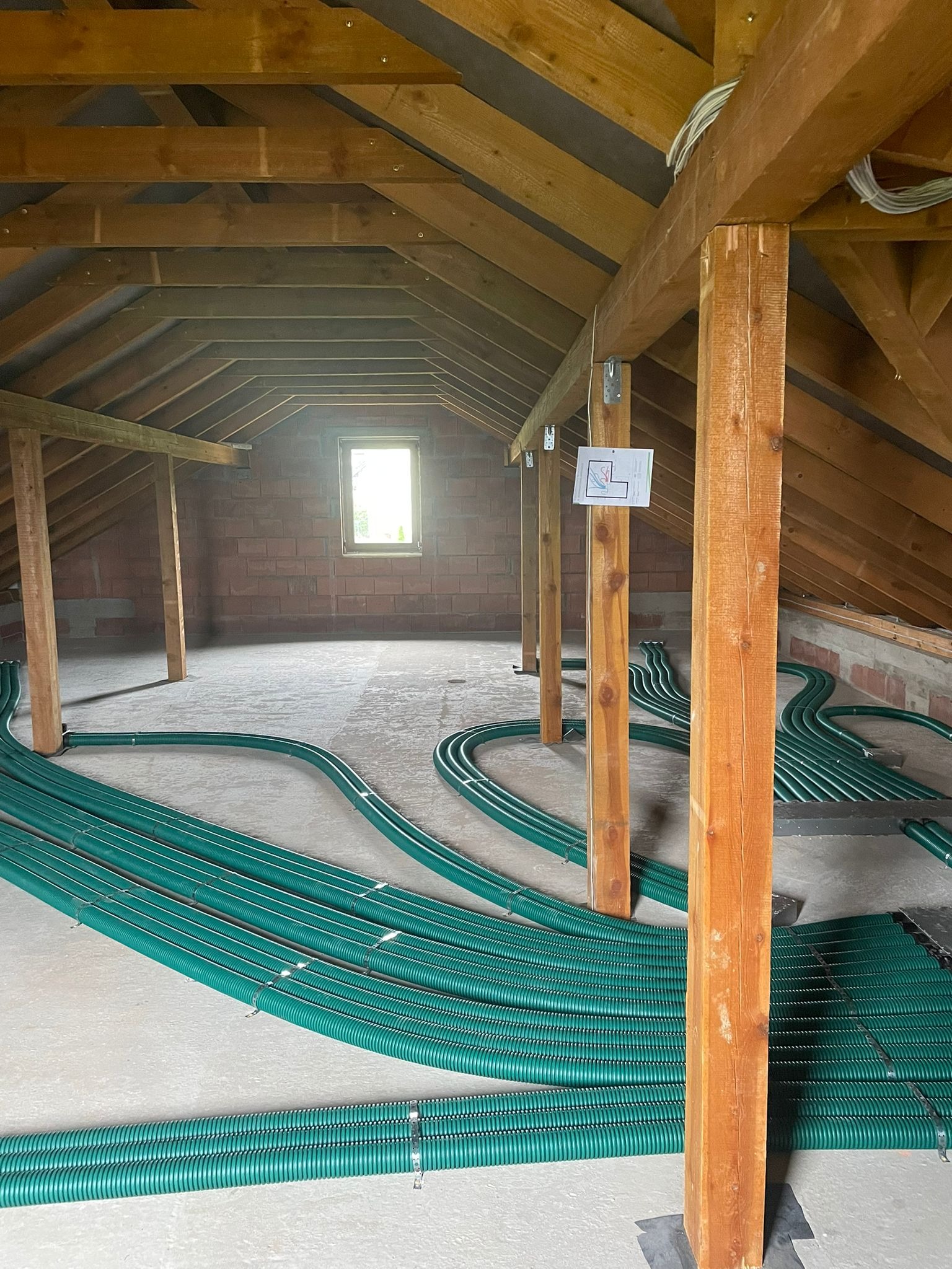 Zielone rury wentylacyjne ułożone równolegle na poddaszu z drewnianą konstrukcją. Widoczne okno i fragment ściany z cegły. Instalacja w trakcie montażu.