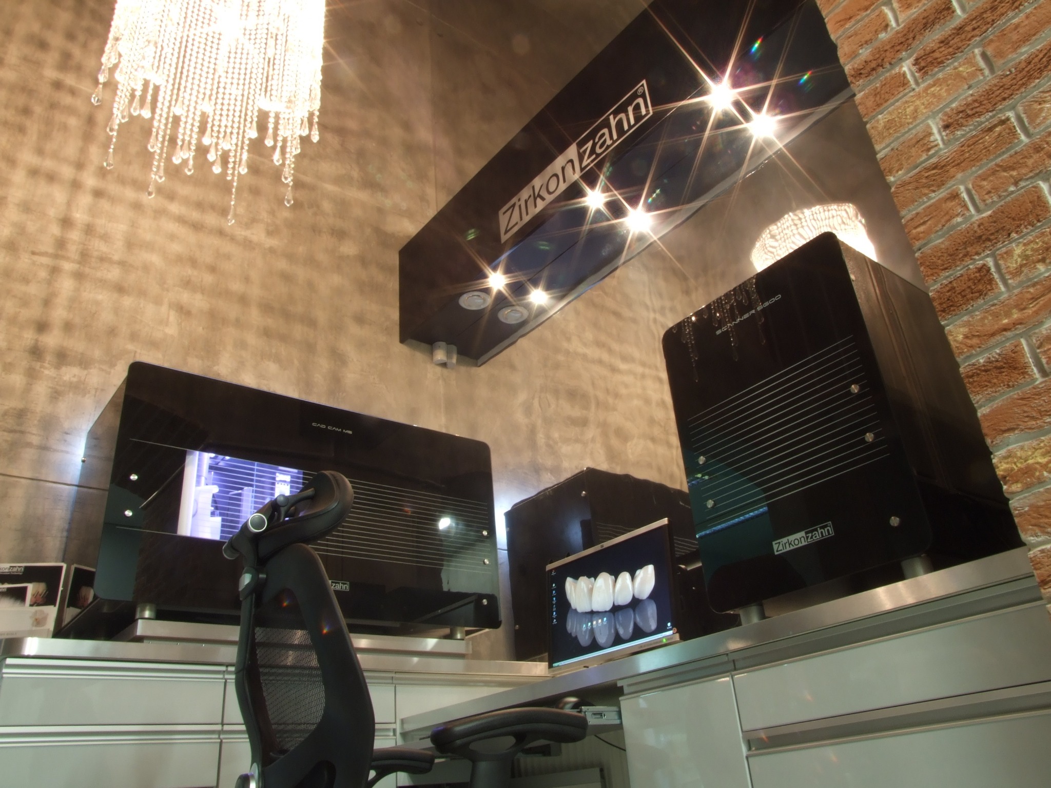 Nowoczesne laboratorium dentystyczne z urządzeniami Zirkonzahn, w tle ściana z cegły i kryształowy żyrandol.