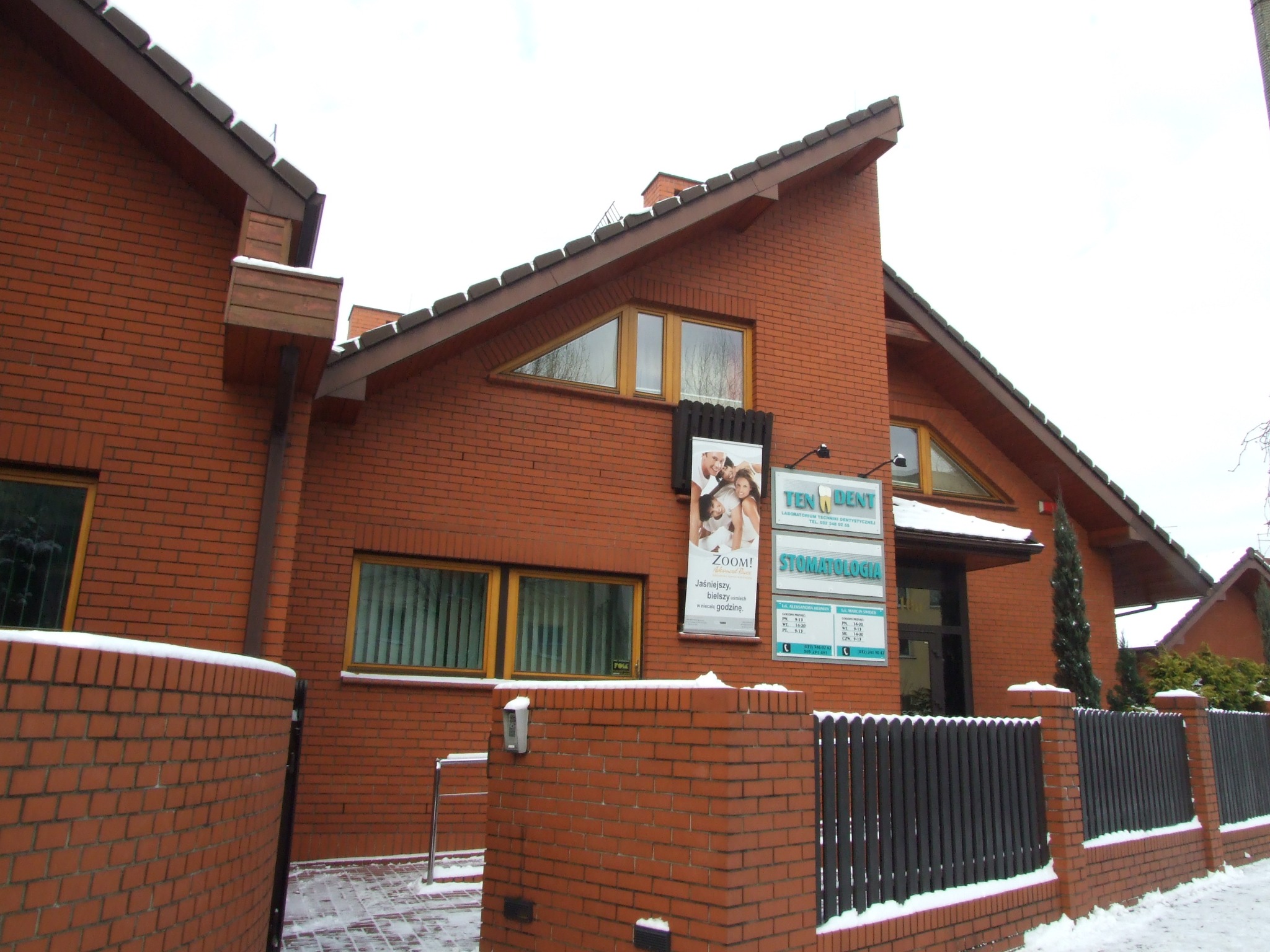 Zewnętrzny widok budynku z czerwonej cegły, w którym mieści się gabinet stomatologiczny Ten-Dent, z reklamą Zoom! na oknie, w zimowej scenerii.