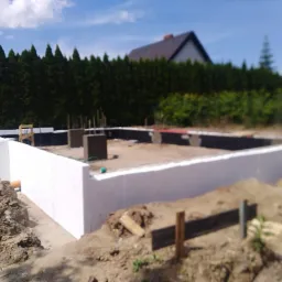 Wykopane fundamenty pod budowę domu, zabezpieczone białym styropianem, widoczne zbrojenia i betonowe słupki, w tle zielony żywopłot i dach domu.