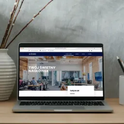 Laptop wyświetla projekt strony internetowej z nowoczesnym biurem w tle, obok wazon z gałązkami i pojemnik z kredkami.