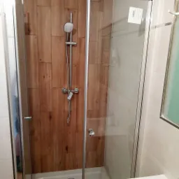 Kabina prysznicowa z chromowaną armaturą i panelem imitującym drewno na ścianie, widoczna naklejka informacyjna na szybie.