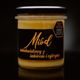 Słoik z jasnym, wielokwiatowym nektarem z imbirem i cytryną, etykieta z nazwą producenta i danymi teleadresowymi, czarne tło z odbiciem.