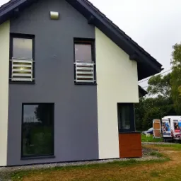 Dwukolorowa elewacja domu jednorodzinnego z widocznymi metalowymi barierkami okiennymi, częściowo ocieplona, z brygadą i samochodem dostawczym w tle.