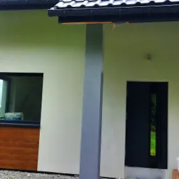 Fragment elewacji domu jednorodzinnego po tynkowaniu, z widoczną aluminiową drabiną opartą o ścianę, wiadrem z farbą na cegle i elementami wykończenia okien w kolorze drewna.