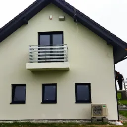 Dwukondygnacyjny dom z jasną elewacją i ciemnoszarą częścią boczną, balkonem oraz klimatyzatorem, widziany z zewnątrz podczas prac wykończeniowych, z pracownikiem na rusztowaniu.