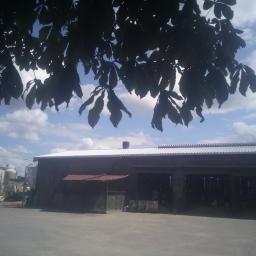 Lux-Dach Józef Bartnicki - Budynek gospodarczy z białym dachem blaszanym, widok spod drzewa z liśćmi, traktor i silosy w tle.