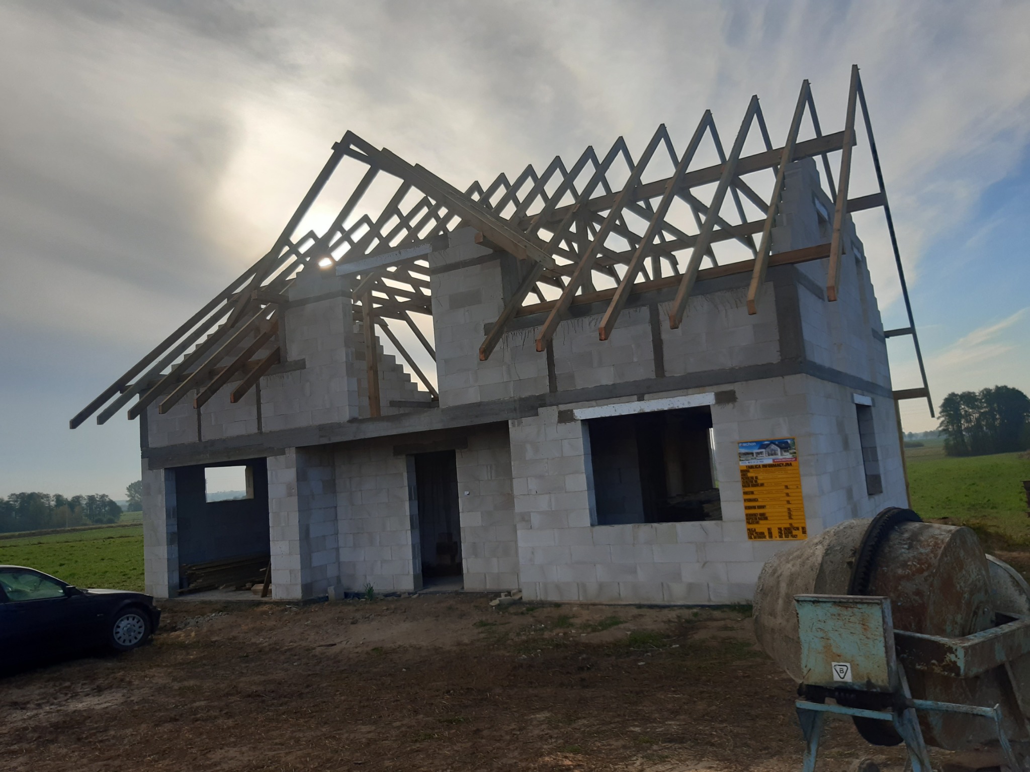 Surowy budynek jednorodzinny w trakcie budowy, ściany z bloczków, widoczna drewniana konstrukcja dachu, betoniarka na pierwszym planie, w tle pole i drzewa.