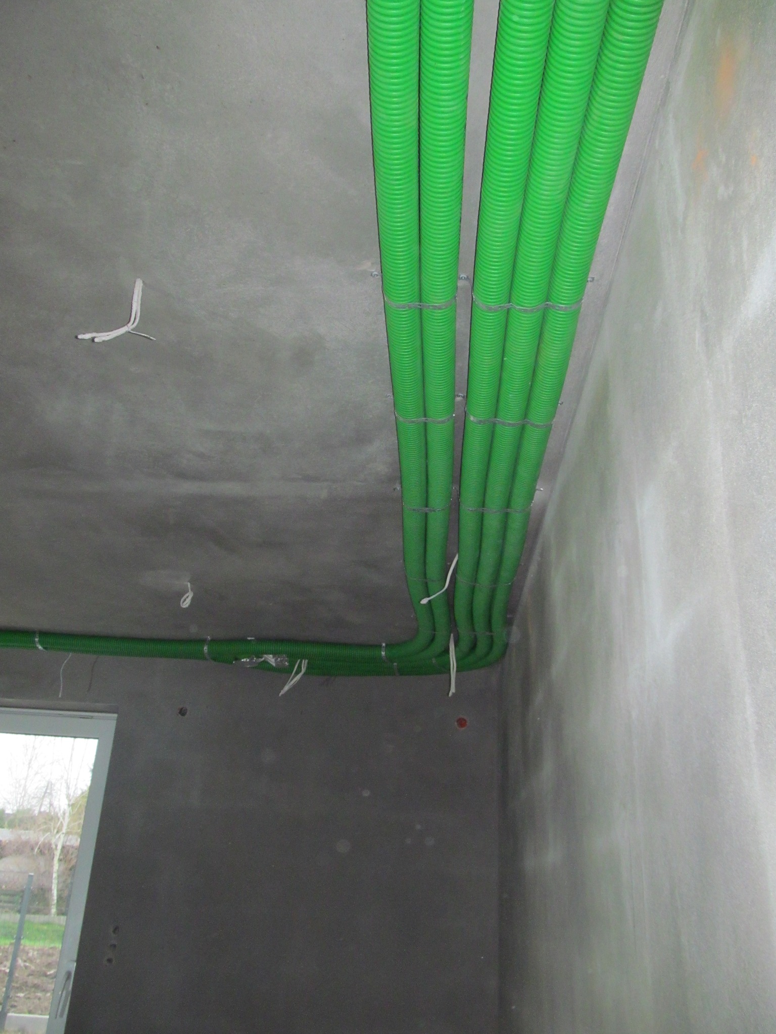 Zielone, karbowane rury wentylacyjne przymocowane do surowego, szarego stropu i ściany w trakcie instalacji, widoczne fragmenty przewodów elektrycznych i okno w tle.