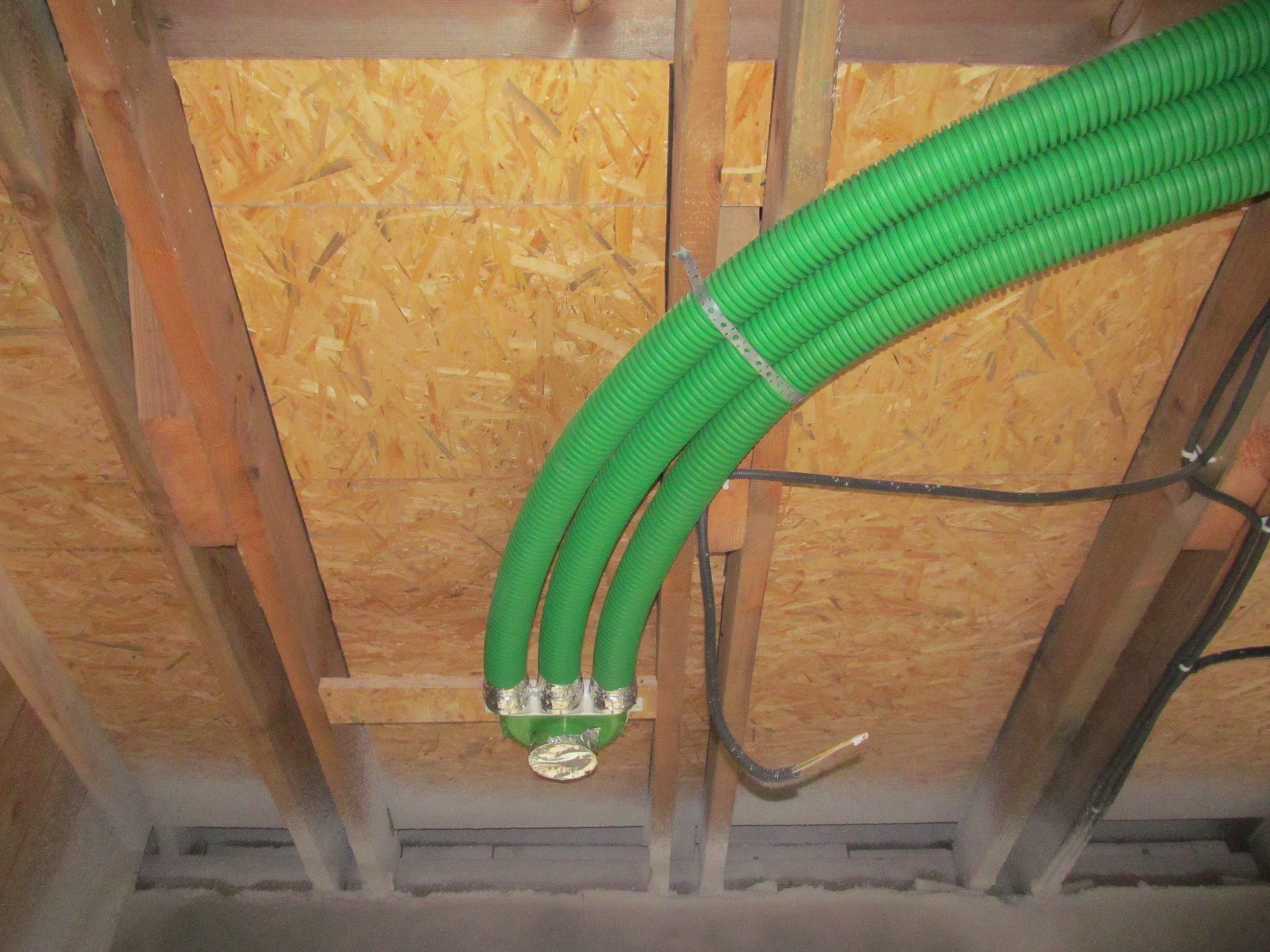 Trzy zielone, karbowane rury wentylacyjne zamocowane do drewnianego stropu z płyt OSB, z widocznymi elementami montażowymi i przewodami elektrycznymi.