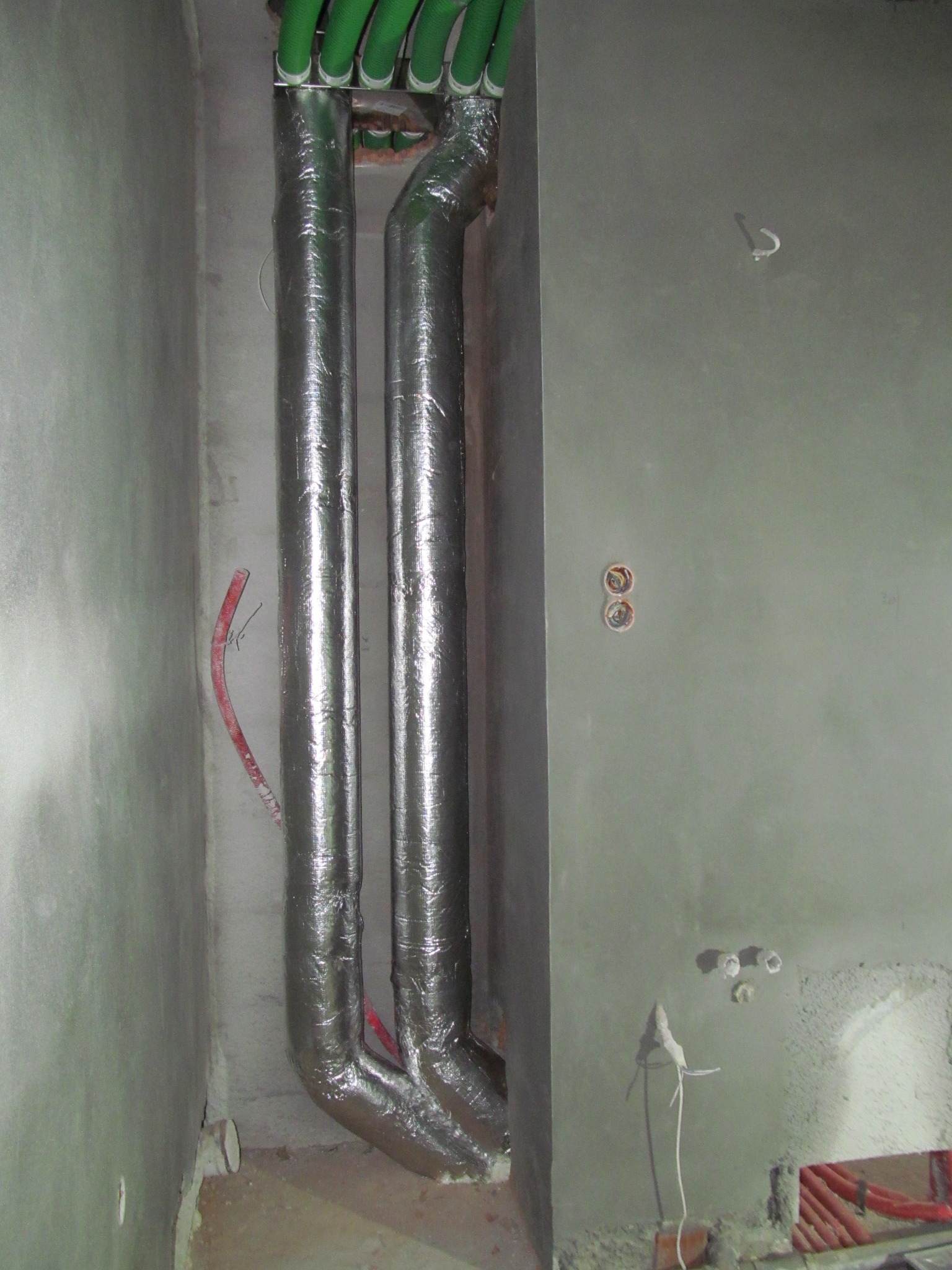 Dwie pionowe rury wentylacyjne w srebrnej izolacji termicznej, z zielonymi rurami u góry, zamontowane w surowym wnętrzu z szarą, nieotynkowaną ścianą.