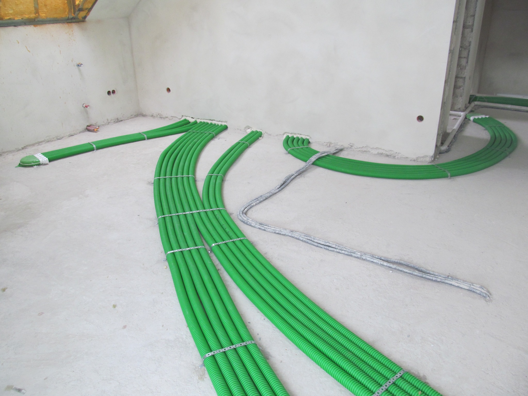 Wnętrze w stanie surowym z ułożonymi wiązkami zielonych, karbowanych rur wentylacyjnych, przymocowanych metalowymi obejmami, oraz szarymi przewodami elektrycznymi, prowadzącymi wzdłuż ścian.