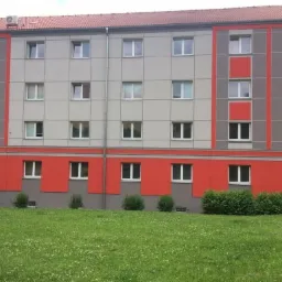 Wielopiętrowy budynek z elewacją w odcieniach szarości i czerwieni, widok z poziomu trawnika.