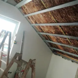 Skośne poddasze w trakcie ocieplania: widoczna wełna mineralna między metalowymi profilami, częściowo obłożone płytami karton-gips, drabina oparta o ścianę, poziomica laserowa.