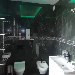 Nowoczesna łazienka z czarnymi marmurowymi płytkami, podświetlanym sufitem LED w kolorze zielonym, białą armaturą (umywalka, toaleta, bidet) i chromowanym grzejnikiem.
