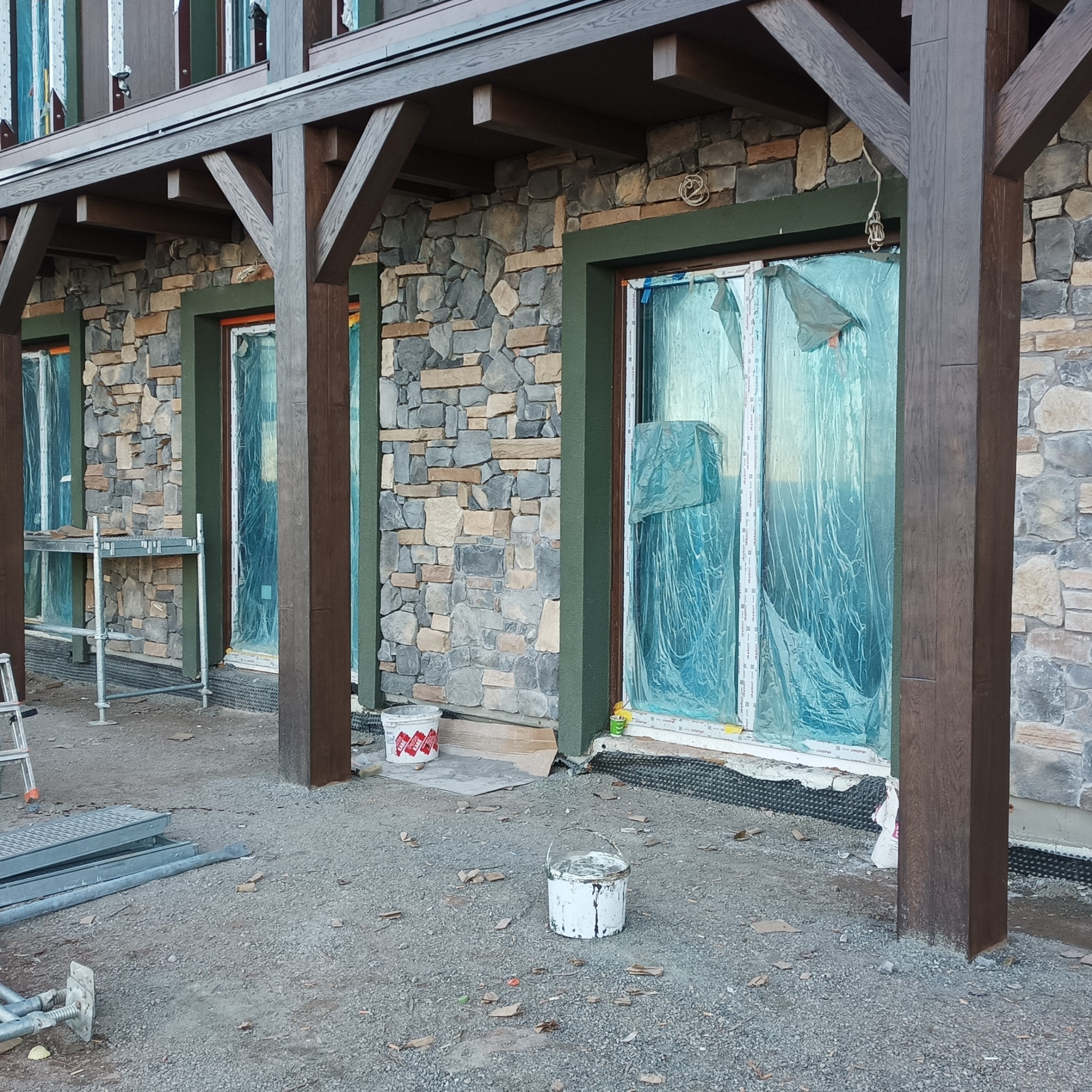 Kamienna elewacja budynku z drewnianymi kolumnami i zabezpieczonymi folią oknami w trakcie prac wykończeniowych, widoczne rusztowanie i wiadra z farbą.