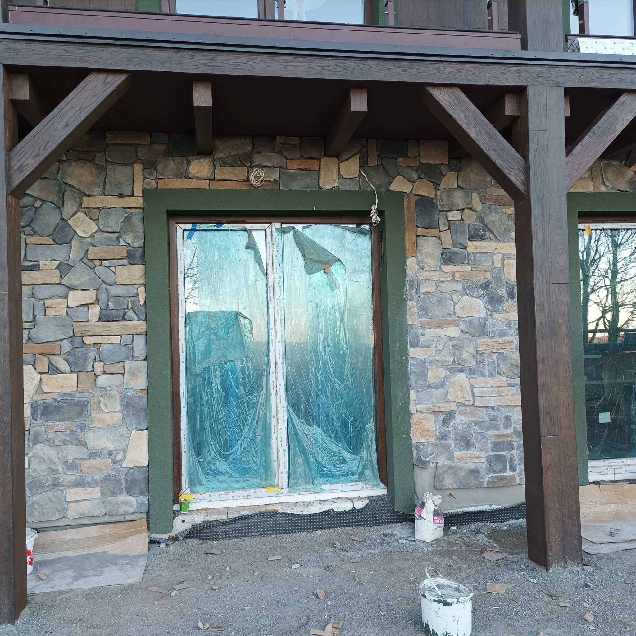 Elewacja budynku w trakcie prac wykończeniowych, widoczne okna zabezpieczone folią, ściana z kamienia i drewniana konstrukcja podtrzymująca dach.