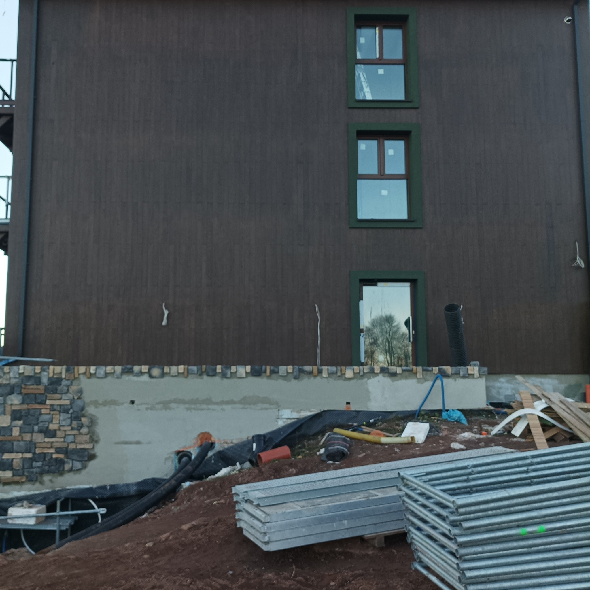 Budynek z brązową elewacją w trakcie wykończenia, widoczne okna z zielonymi ramami, fragment wykończonej kamiennej elewacji na dole, rury, rusztowania i materiały budowlane na pierwszym planie.
