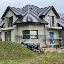 Dom jednorodzinny w trakcie ocieplania elewacji, widoczne rusztowania, częściowo otynkowane ściany, czarna dachówka i materiały budowlane na trawniku.