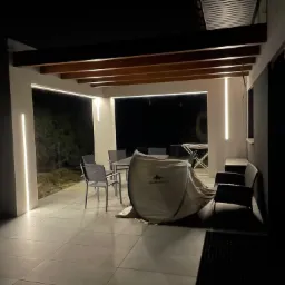 Nowoczesna, oświetlona listwami LED weranda z drewnianym zadaszeniem, wyposażona w meble ogrodowe i rozłożony namiot turystyczny Quechua, widziana nocą.