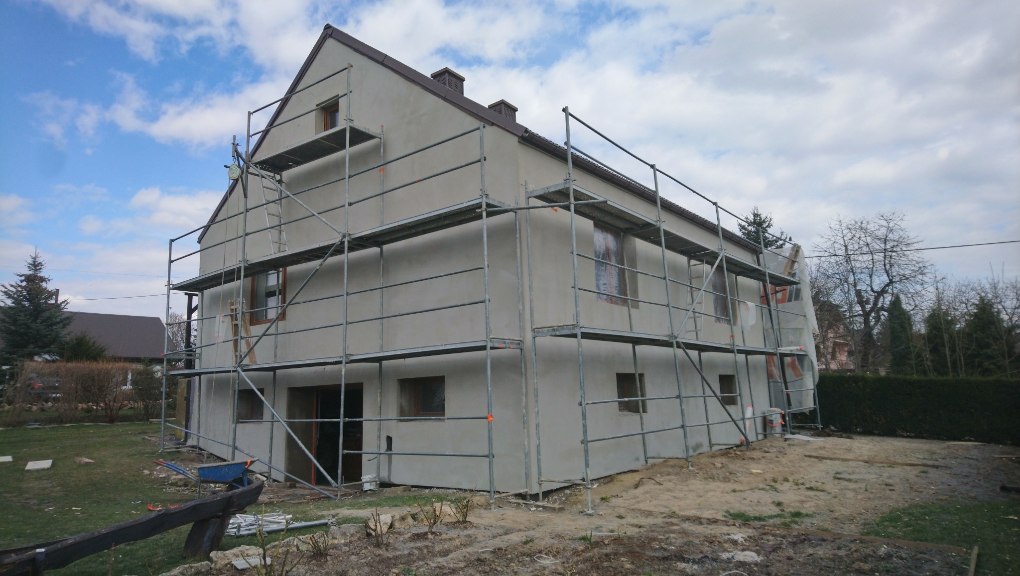 Dwupiętrowy dom w trakcie prac elewacyjnych, otoczony rusztowaniem, z widocznymi oknami zabezpieczonymi folią, na tle pochmurnego nieba i drzew bez liści.