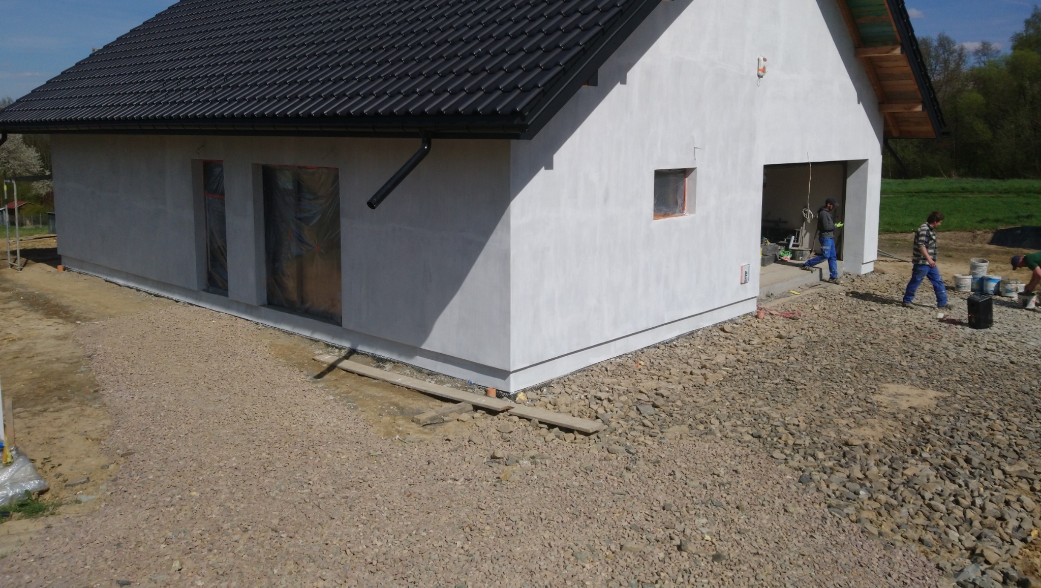 Surowa elewacja domu jednorodzinnego w trakcie prac wykończeniowych, widoczne okna zabezpieczone folią, czarna dachówka, teren wokół wysypany żwirem, pracownicy budowlani w tle.