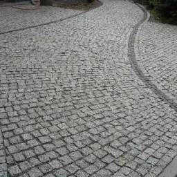 Ścieżka z szarej kostki granitowej ułożona w łuk, obrzeża z ciemniejszej kostki, widok z góry.