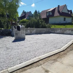 Przygotowany teren pod wylewkę, ogrodzony bloczkami betonowymi i obrzeżami chodnikowymi, z widokiem na domy w tle.