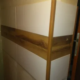 Biała zabudowa meblowa z drewnianymi akcentami, widoczne usłojenie drewna, fragment drzwi po prawej stronie kadru.