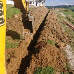 Żółta koparka tworzy wąski wykop w ziemi, w tle dom w stanie surowym zamkniętym i pagórkowaty krajobraz.