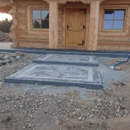 Dwa stopnie z kostki brukowej prowadzące do drewnianego domu, widoczna ubijarka w tle, teren częściowo utwardzony