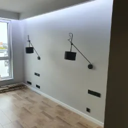 Północno-zachodnie ujęcie wnętrza pokoju z drewnopodobną podłogą, szarymi ścianami, czarnymi lampami ściennymi i gniazdkami elektrycznymi; widoczne paczki paneli podłogowych przy ścianie