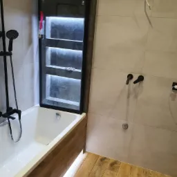 Nowoczesna łazienka z wanną obudowaną drewnopodobnymi panelami, czarną armaturą i podświetlaną wnęką w ścianie. Na podłodze i ścianach płytki w odcieniach beżu i szarości. Na podłodze leży tuba...