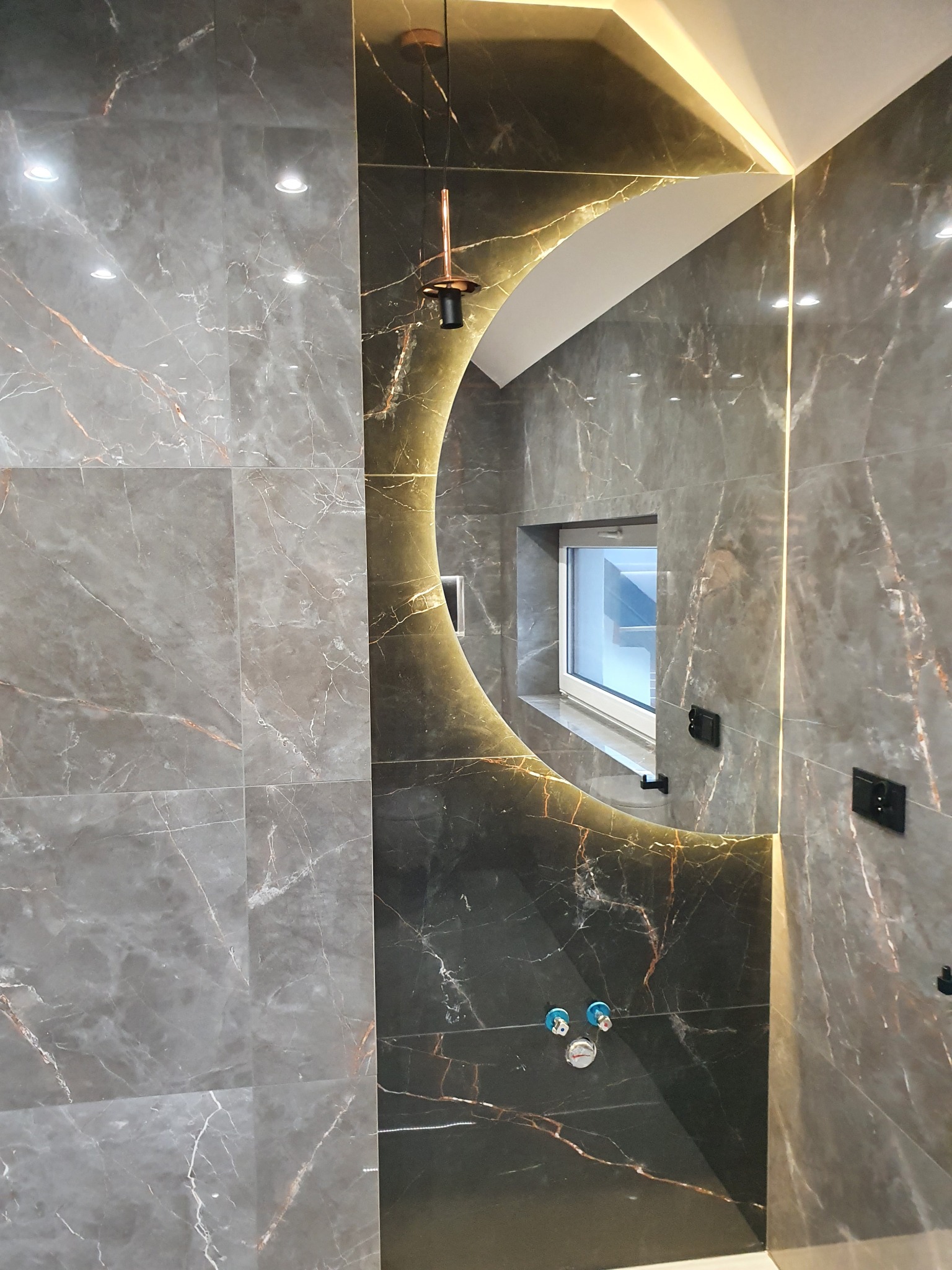 Wykończona łazienka z szarymi płytkami imitującymi marmur, okrągłe podświetlane lustro, nowoczesna lampa wisząca i okno z widokiem na schody.