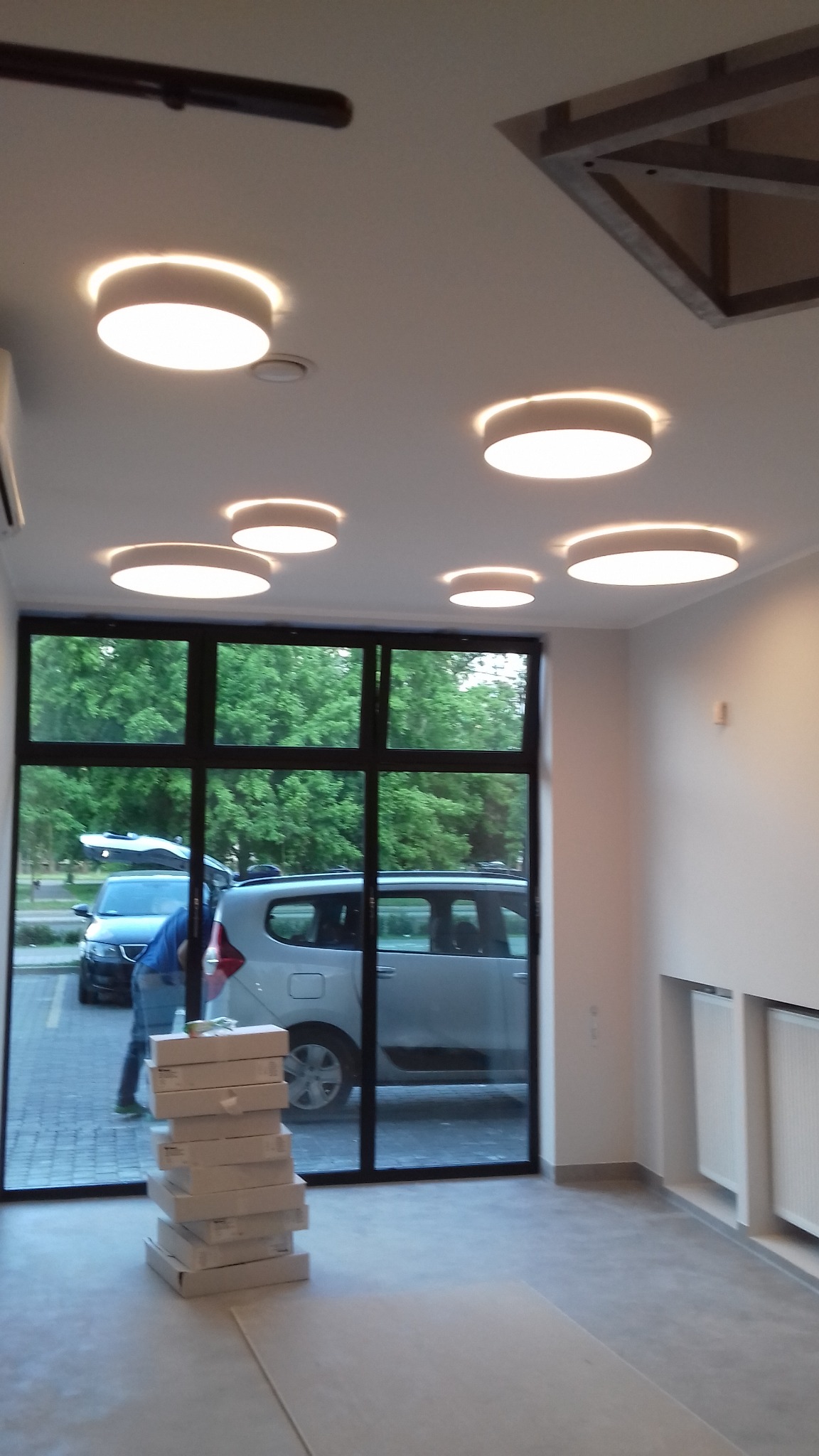 Wnętrze w trakcie wykańczania: ułożona podłoga, stos pudełek z panelami, zamontowane okrągłe lampy sufitowe, widok na zewnątrz przez duże okno.