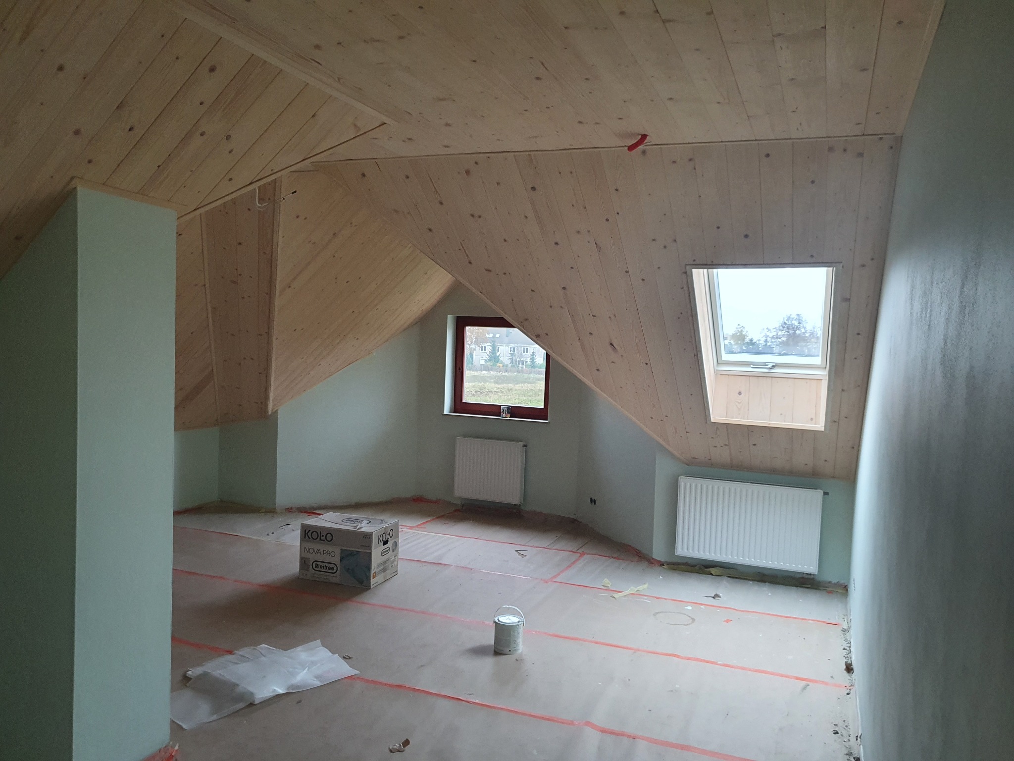 Pokój na poddaszu w trakcie prac wykończeniowych: drewniana boazeria na skośnym suficie, pomalowane na jasnozielono ściany, zabezpieczona folią podłoga, widoczny grzejnik i okno dachowe.